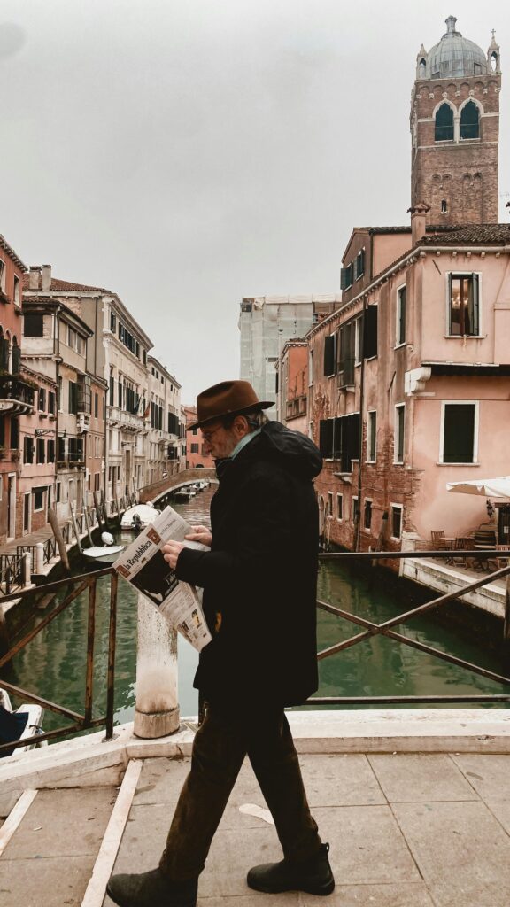 gentleman in a fine hat and coat reading an italian newspaper as he crosses a canal bridge in venice